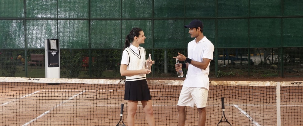 Two tennis players talking over Boon glass bottles at the net, a Boon purifier courtside